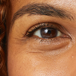 A detailed close-up of a person's left eye area, highlighting the natural signs of aging. Visible crow's feet and fine lines crinkle outward from the corner of the eye and underneath the lower eyelid against a warm-toned complexion.