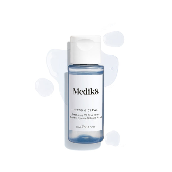 A travel-size bottle (30ml) of Medik8 Press & Clear tonic with a white flip-top cap, surrounded by clear blue droplets on a white surface. Text identifies it as an  Exfoliating 2% BHA Tonic. -25