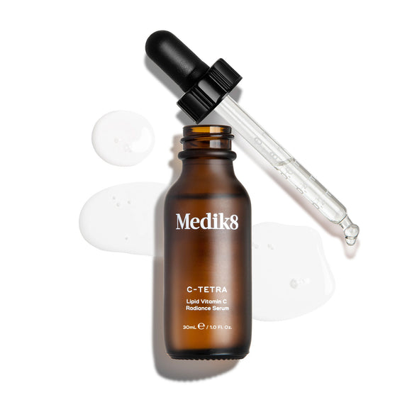 An overhead "packshot" of a 30ml bottle of Medik8 C-Tetra Lipid Vitamin C Radiance Serum. The amber glass bottle is uncapped and stands centered, with its black dropper lid resting diagonally behind it. A clear, liquid serum is artistically splashed and pooled on the clean white background around the bottle, with a few droplets visible on the glass pipette. The lighting is bright, casting soft shadows to the left and creating a clinical yet elegant aesthetic.-5
