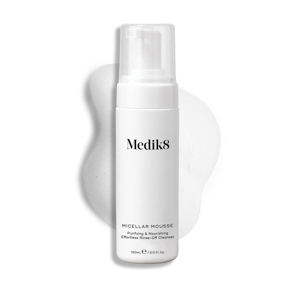 Medik8 Micellar Mousse pump bottle displayed on a white background with a smear of white foam behind it, highlighting the product's texture.-1