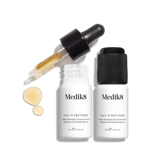 Two small white bottles of Oxy-R Peptides serum are displayed. A black dropper tool lies nearby, and two drops of yellow serum are visible on the white surface-17