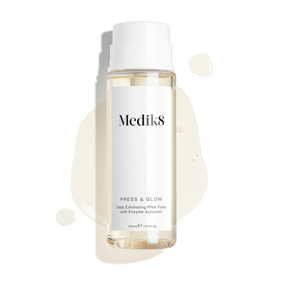 Medik8 Press & Glow tonic bottle stands against a clean white background with translucent liquid droplets and a puddle, highlighting its watery texture. Text identifies the product as a 200mL  Daily Exfoliating PHA Tonic with Enzyme Activator. -1