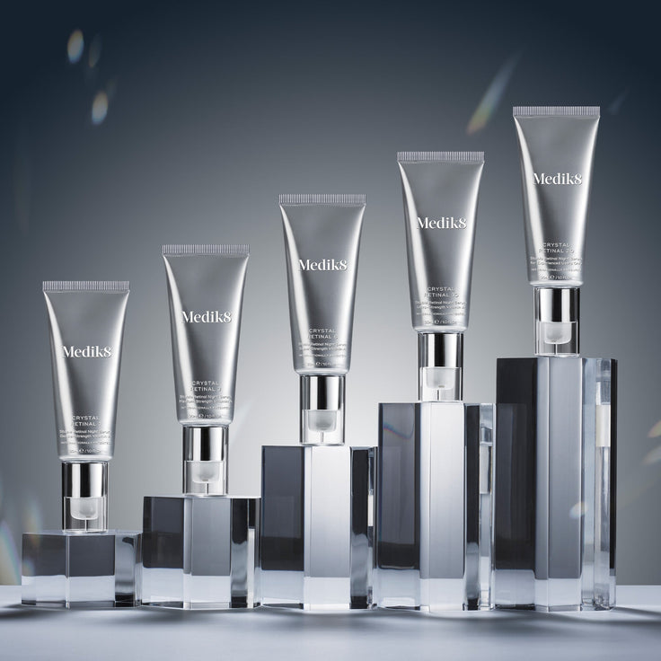 Six metallic silver tubes of Medik8 Crystal Retinal serum arranged in a rising staircase formation from left to right. Each tube stands vertically on a clear, reflective glass block of increasing height. The tubes display escalating product strengths: Crystal Retinal 1, 3, 6, 10, 20, and 24 against a solid gradient grey background.