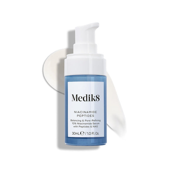 Medik8 Niacinamide Peptides serum bottle with a white pump top, resting on a white surface with a smear of white serum behind it.-4