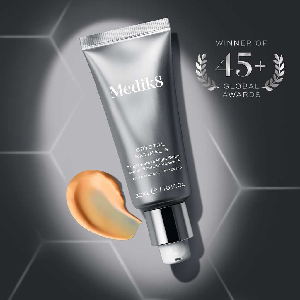 A metallic silver tube of Medik8 Crystal Retinal 6 serum angled diagonally within a bright spotlight on a dark grey background patterned with faint geometric hexagons. Next to the tube pump is a thick, curved swatch of peach-colored cream. In the top right corner, a silver metallic laurel wreath emblem reads "WINNER OF 45+ GLOBAL AWARDS".