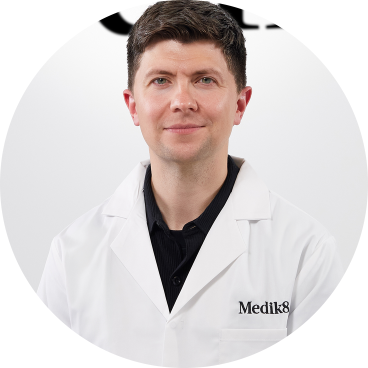 A headshot of Daniel Isaacs, Chief Product Officer of Medik8. He is wearing a black collared shirt underneath a white professional lab coat embroidered with the black "Medik8" logo on the left pocket. The photo is cropped into a perfect circle with a bright white background.
