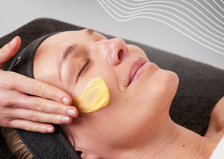 A close-up view of a person lying down with closed eyes during a professional facial treatment. A thick layer of yellow skincare product is smoothly spread across their left cheek while a practitioner's hands gently frame their face. The top right corner features abstract, thin white wavy lines over a grey background.