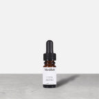 A travel-sized 8mL amber glass bottle of Medik8 C-Tetra lipid vitamin C radiance serum standing next to its white cardboard carton. The products are displayed on a textured grey surface against a clean white background