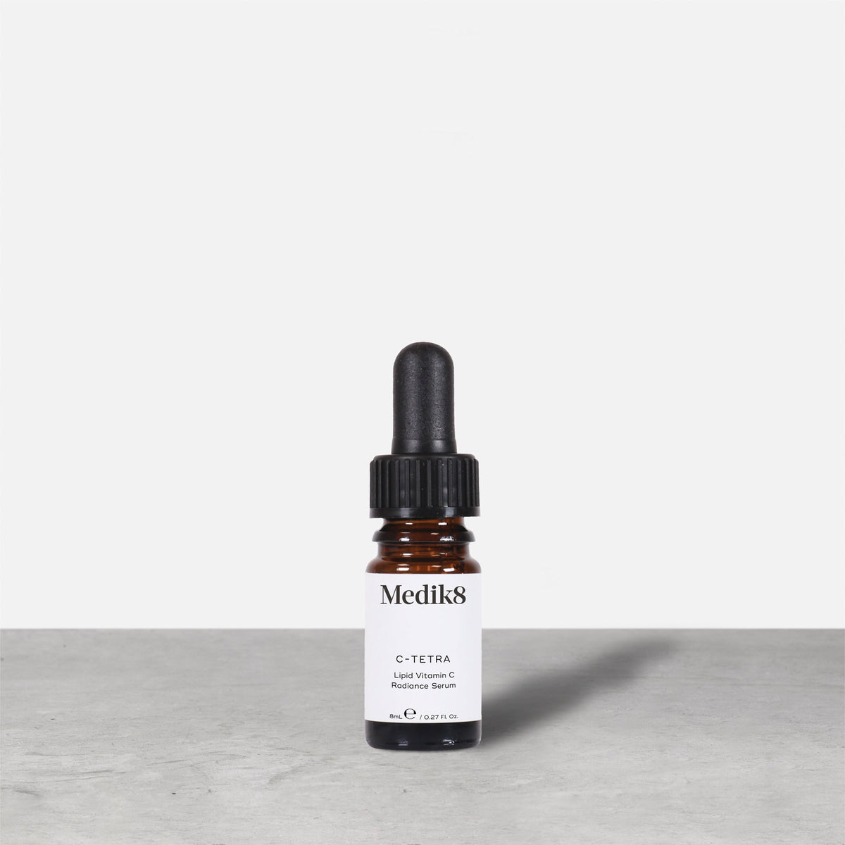 A travel-sized 8mL amber glass bottle of Medik8 C-Tetra lipid vitamin C radiance serum standing next to its white cardboard carton. The products are displayed on a textured grey surface against a clean white background