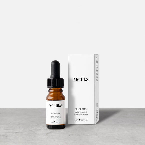 A travel-sized 8mL amber glass bottle of Medik8 C-Tetra lipid vitamin C radiance serum standing next to its white cardboard carton. The products are displayed on a textured grey surface against a clean white background.-12