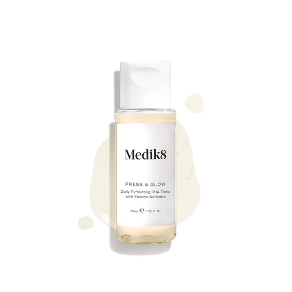A 30mL travel-sized clear bottle of Medik8 Press & Glow tonic with a white cap, displayed on a clean white background. The bottle rests on a puddle of the pale yellow liquid with scattered droplets around it, highlighting its lightweight texture. Text on the label reads ""Daily Exfoliating PHA Tonic with Enzyme Activator.