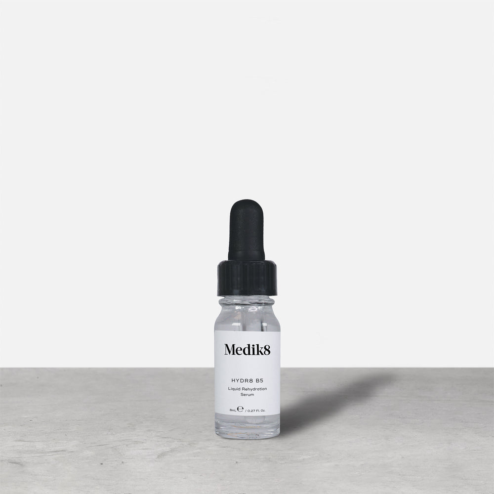 Medik8 Hydr8 B5 travel-sized serum bottle stand on a grey textured surface against a neutral background. Text on the 8mL bottle identifies it as a "Liquid Rehydration Serum.
