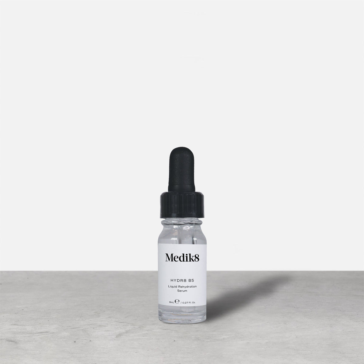 Medik8 Hydr8 B5 travel-sized serum bottle stand on a grey textured surface against a neutral background. Text on the 8mL bottle identifies it as a "Liquid Rehydration Serum.