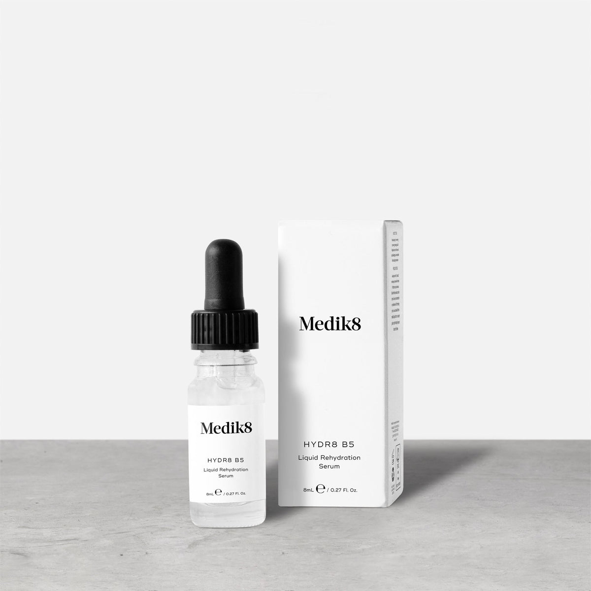 Medik8 Hydr8 B5 travel-sized serum bottle stands next to its matching white product carton on a grey textured surface against a neutral background. Text on the 8mL bottle identifies it as a ""Liquid Rehydration Serum."""