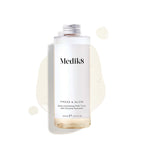 A 200mL clear refill bottle of Medik8 Press & Glow exfoliating tonic featuring a silver screw cap. Set against a white background, the bottle sits amidst a puddle and droplets of the pale yellow tonic, showcasing its liquid texture. The label text reads ""Daily Exfoliating PHA Tonic with Enzyme Activator."