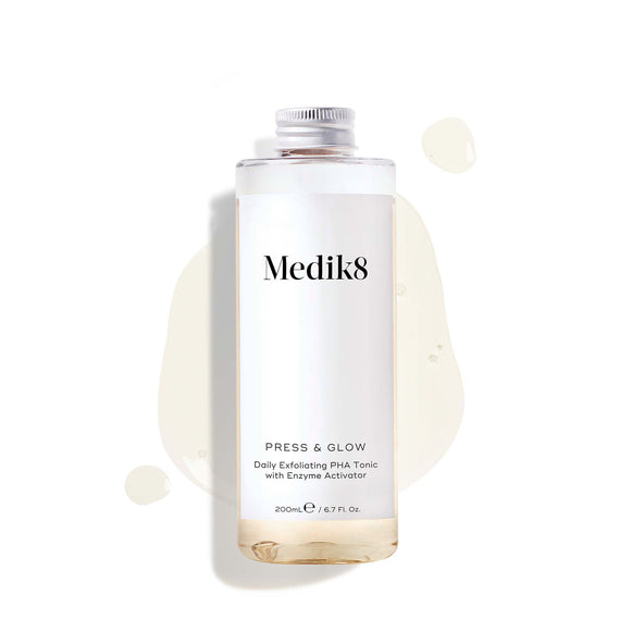 A 200mL clear refill bottle of Medik8 Press & Glow exfoliating tonic featuring a silver screw cap. Set against a white background, the bottle sits amidst a puddle and droplets of the pale yellow tonic, showcasing its liquid texture. The label text reads ""Daily Exfoliating PHA Tonic with Enzyme Activator."""